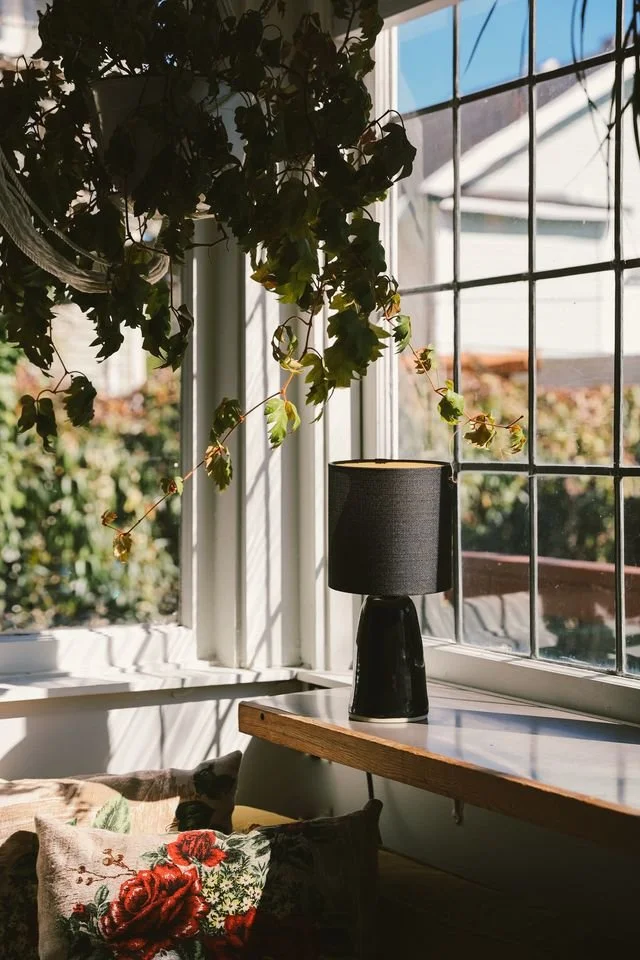 Sunlight streams into a cozy corner with a large window, a black table lamp made by Natalya Jane sits on a wooden ledge, and green vines hanging from above, creating a warm, inviting atmosphere.