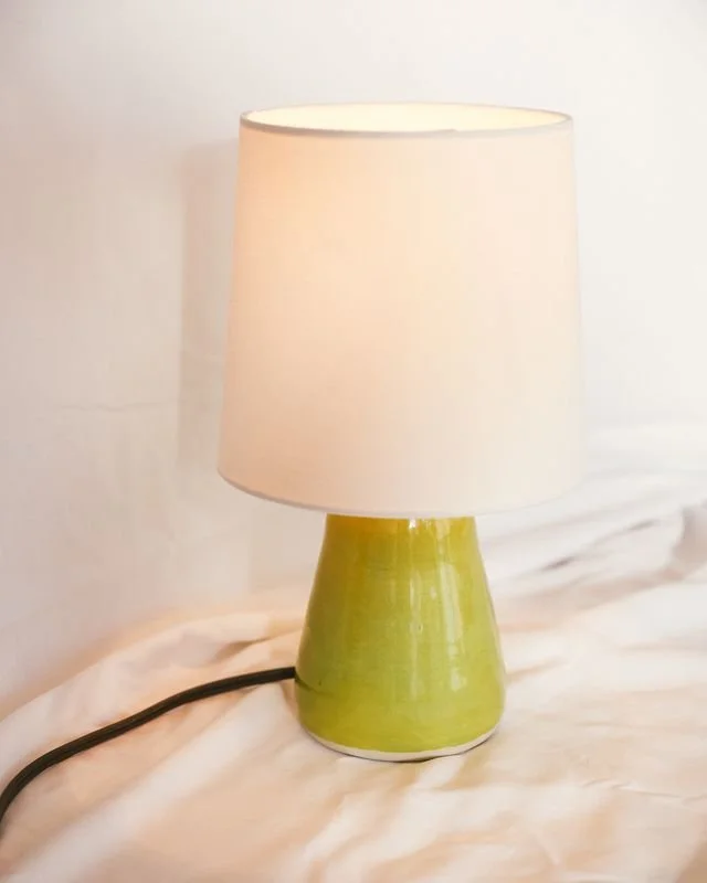 A Green ceramic table lamp with a white lampshade, made by Natalya Jane, on a white surface against a white wall.