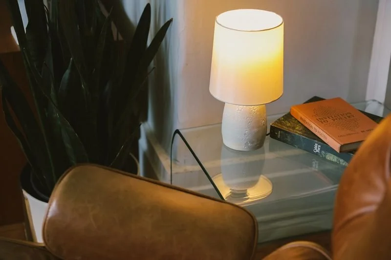 A cozy corner with a glass-top table, a small white table lamp made by Natalya Jane, a stack of books, and a leather chair with a rounded back.