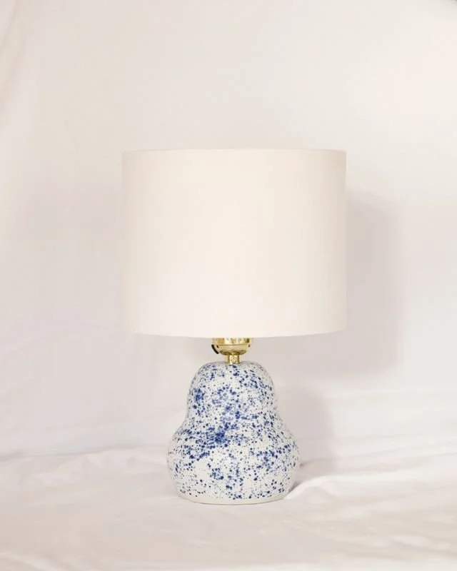 A table lamp with a blue and white speckled ceramic base and a white lampshade made by Natalya Jane, on a white surface against a white wall.