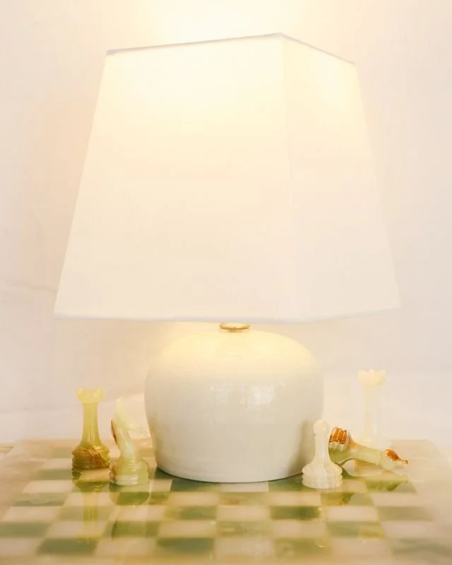 A chess set with chess pieces on a table, illuminated by a table lamp with a white shade made by Natalya Jane.