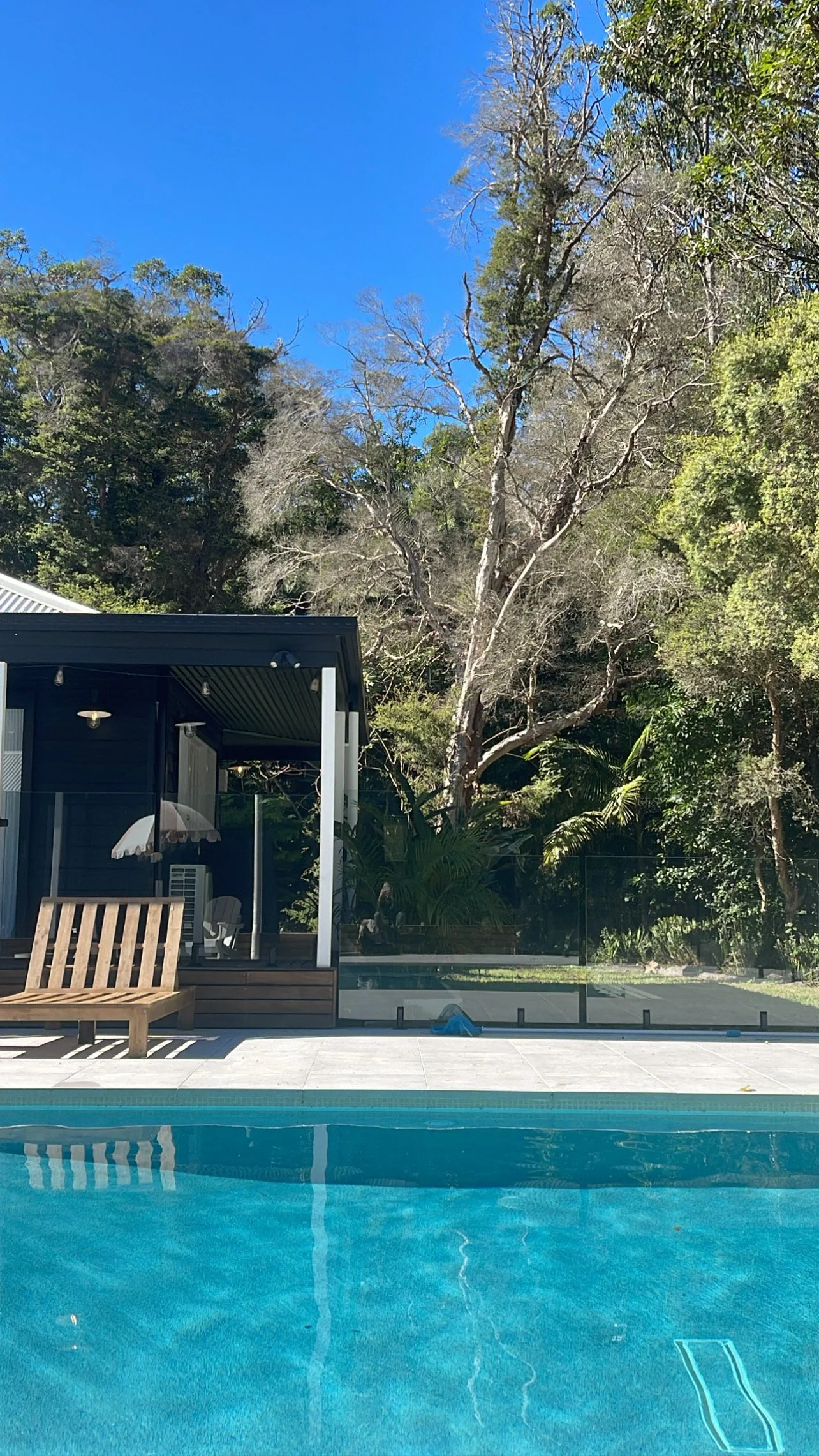View of a backyard with a swimming pool, a wooden bench, and a small patio area with an outdoor seating area, surrounded by tall trees and greenery, under a bright blue sky.