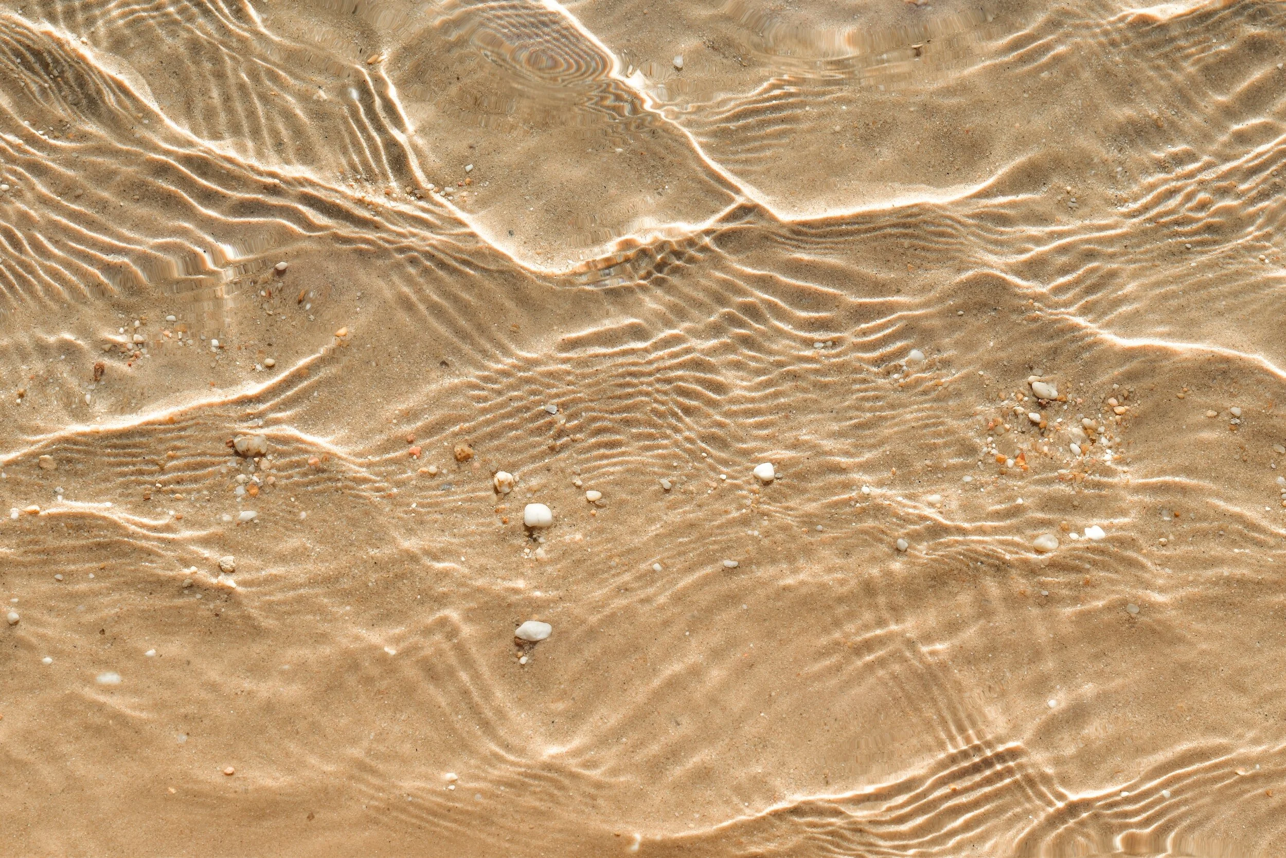 Close-up of sandy beach with shallow, clear water creating ripples and small shells scattered on the sand.