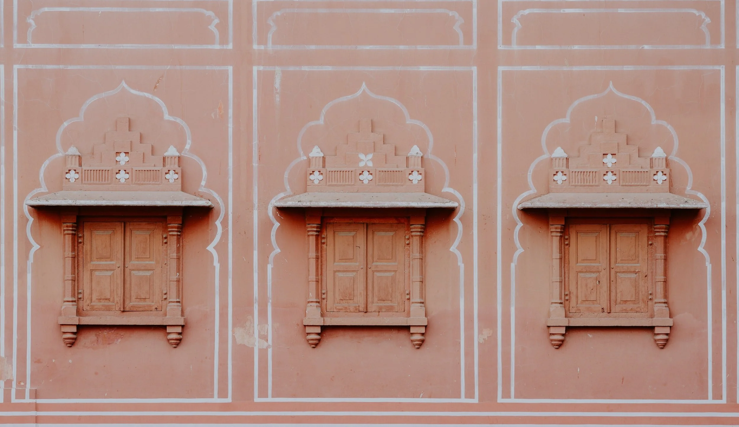 Three decorative windows or niches with closed wooden shutters on a peach-colored wall, featuring intricate architectural details.