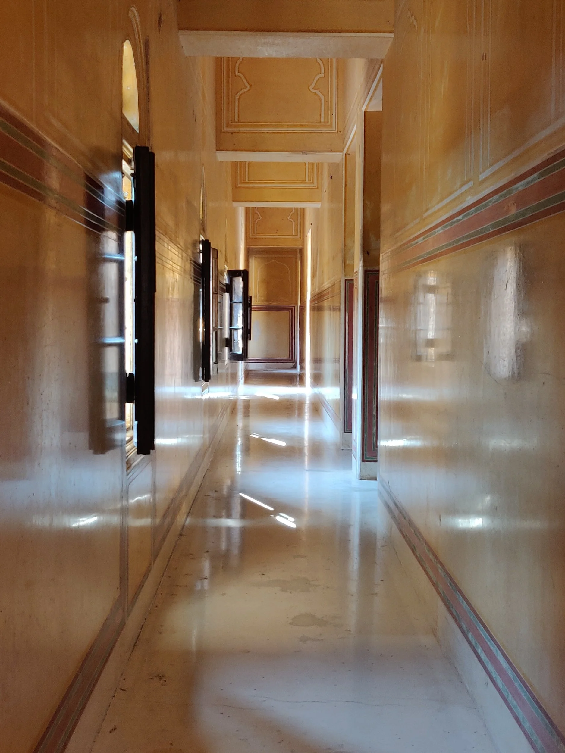 A long, narrow corridor with yellow-golden walls and decorative ceilings. Windows on one side allow sunlight to create reflections on the polished floor, illuminating the hallway.