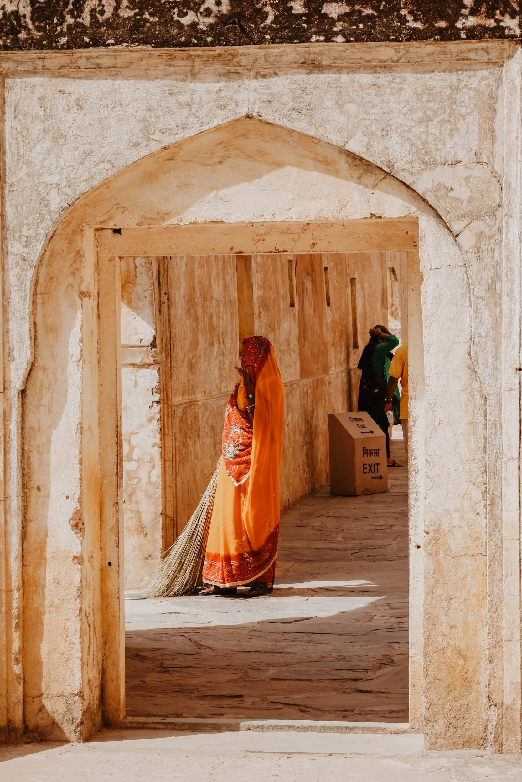 A woman dressed in a traditional orange sari sweeps the ground with a broom inside an ancient, weathered stone doorway. Two other people are visible in the background, one touching their head and the other standing near an exit sign in a historical structure.