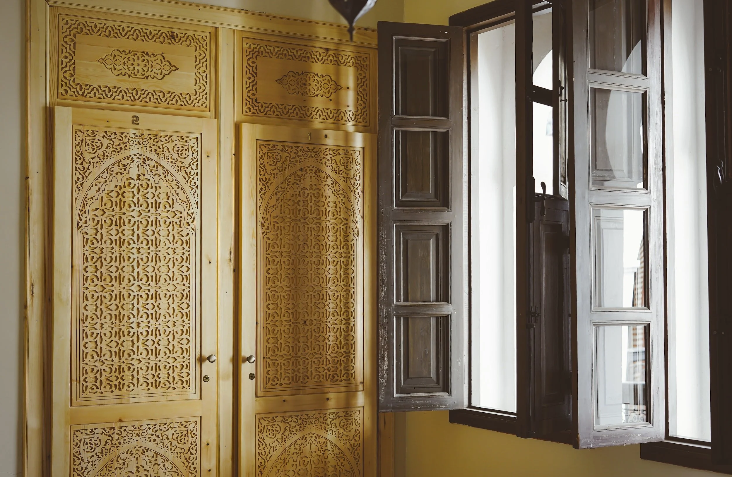 Wooden louvered window open to the outside, next to a decorative wooden wardrobe with intricate carvings.