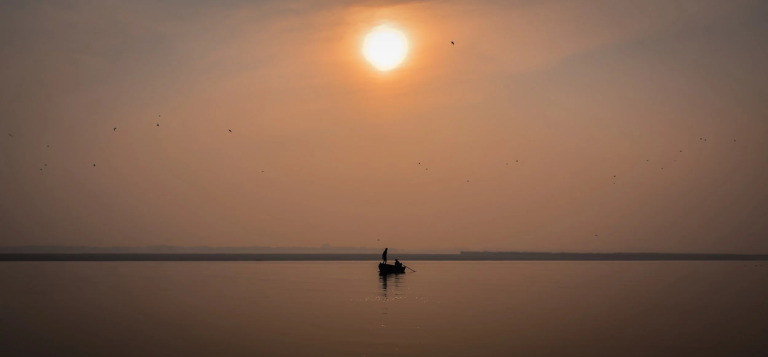 A boat with two people on a calm body of water during sunset or sunrise, with the sun low in the sky and several birds flying overhead.