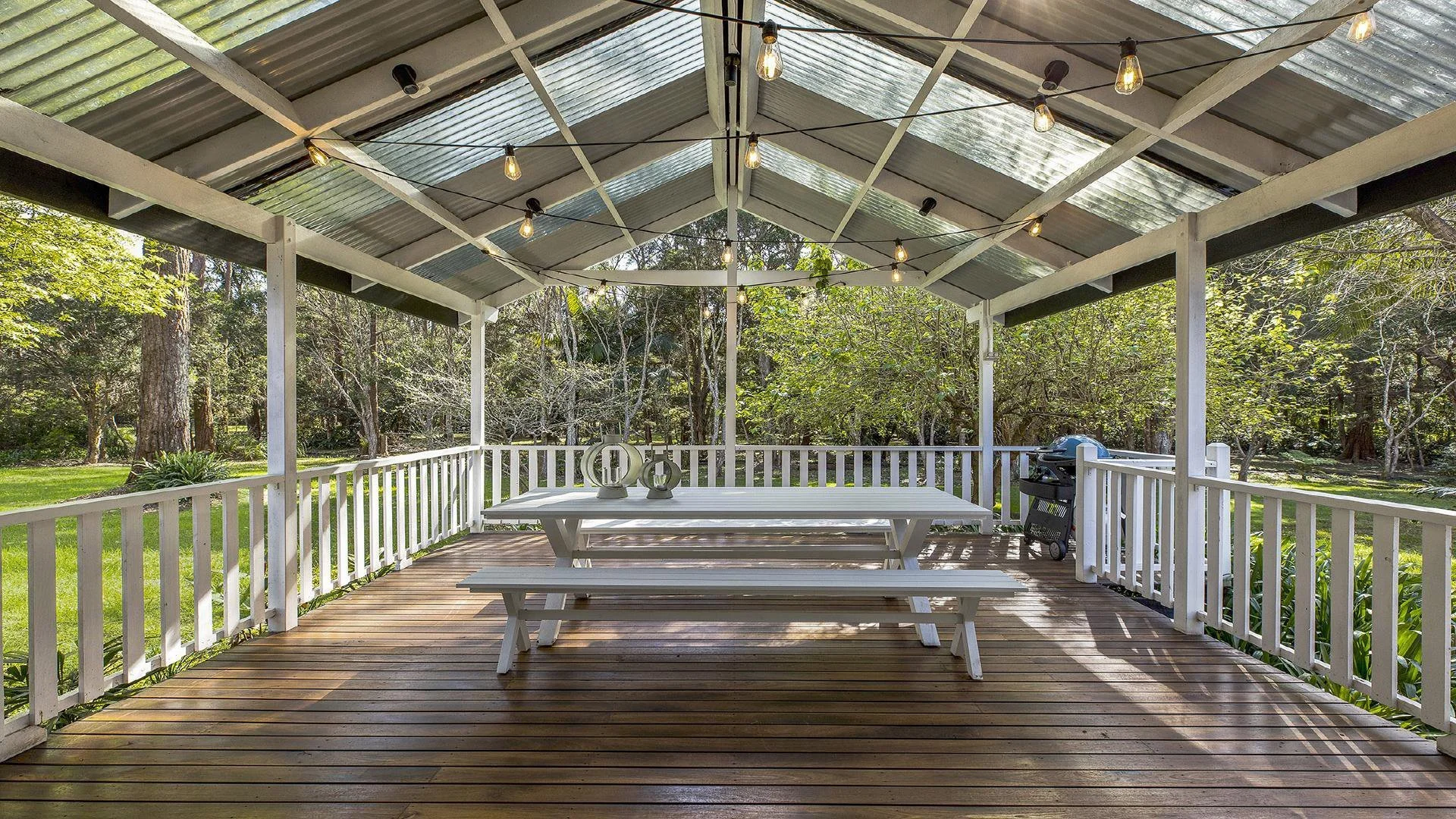 Covered outdoor deck with wooden flooring, white railing, picnic table with benches, string lights hanging from roof, and a grill in the corner, surrounded by greenery and trees.