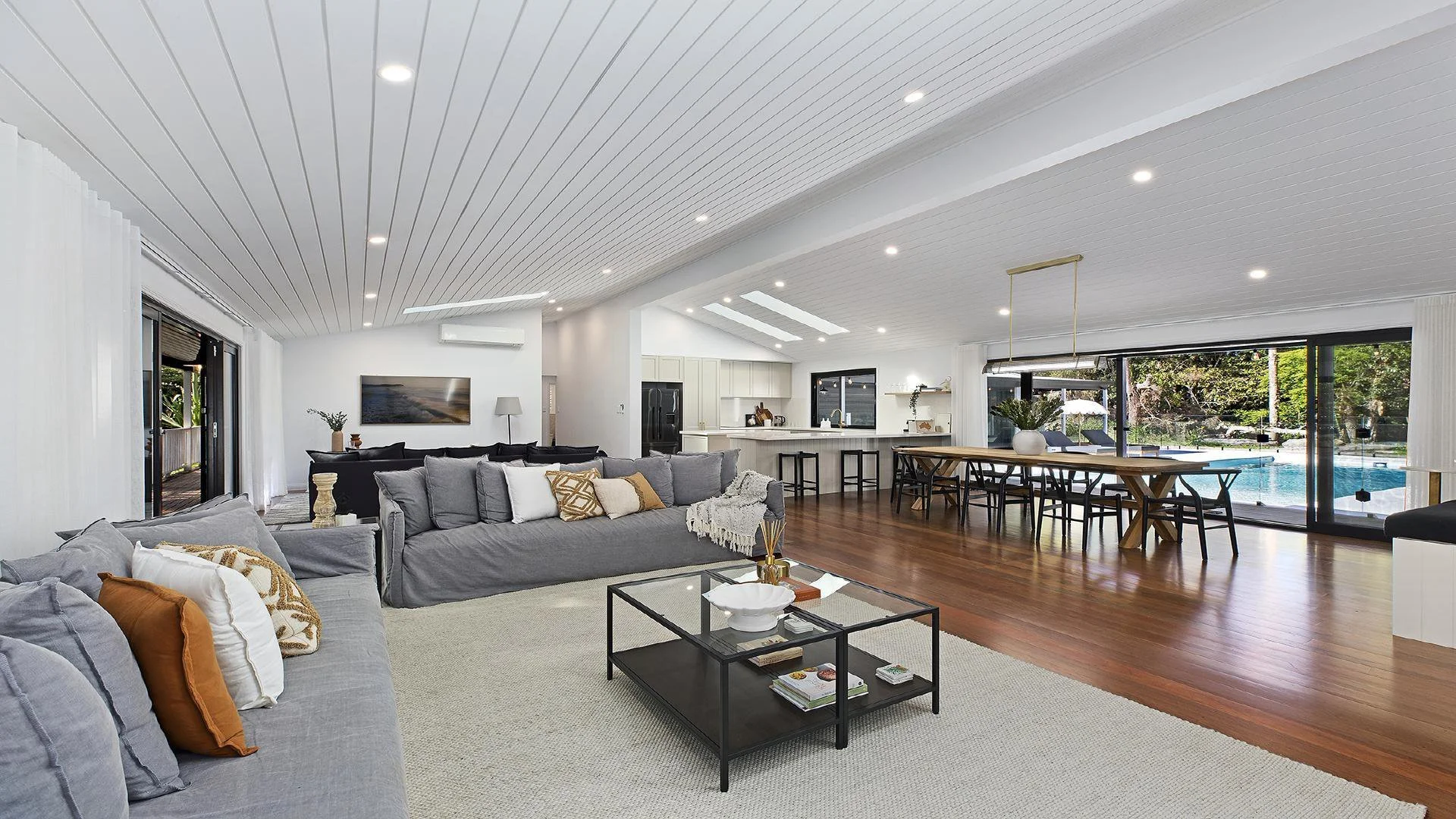 Open-concept living room and dining area with large windows, hardwood floors, modern furniture, and a view of the backyard pool.