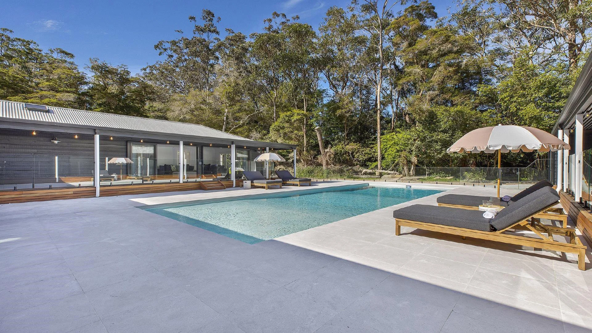 Modern house with outdoor pool area, lounge chairs with umbrellas, surrounded by trees and blue sky.