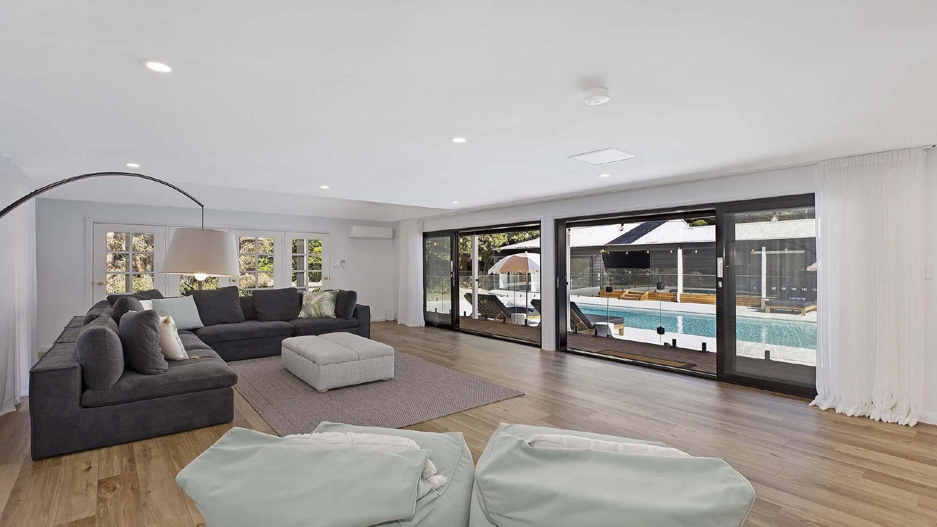 Living room with gray sofas, pillows, a beige ottoman, large sliding glass doors leading to a pool area, with wooden floors, white walls, and curtains