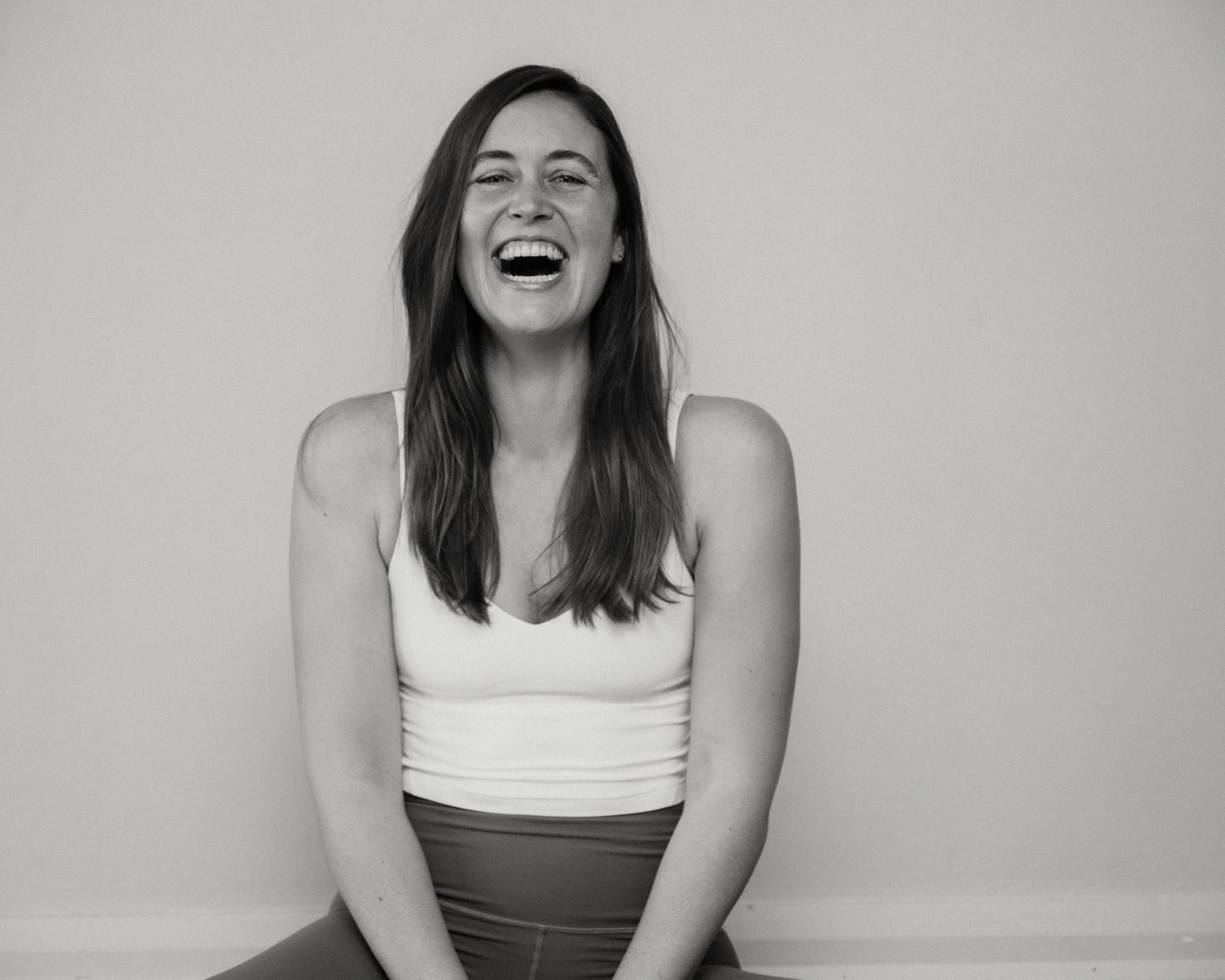 Black and white photo of a woman with long hair, smiling and laughing, wearing a sleeveless top and sitting against a plain background.
