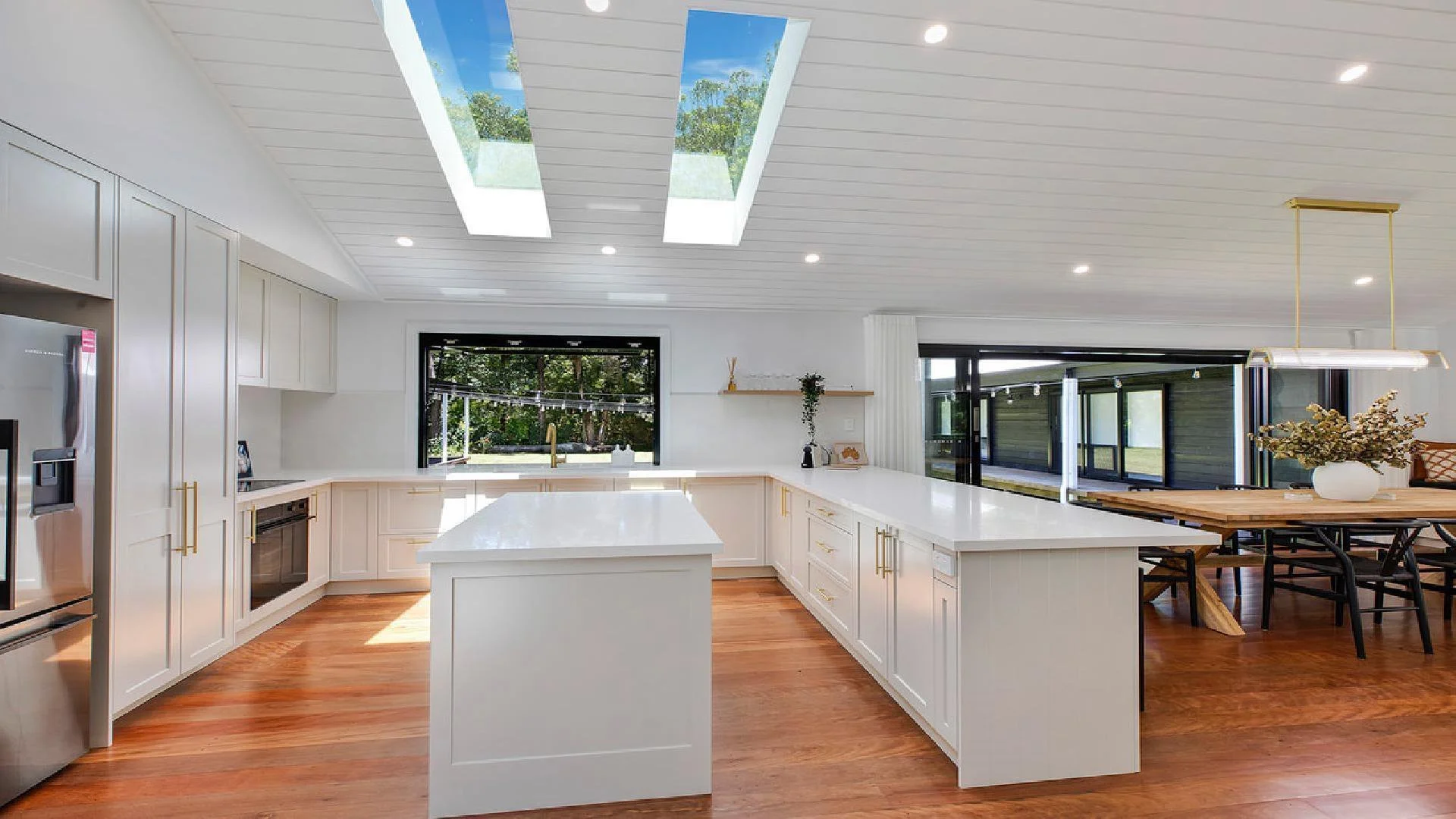Bright modern kitchen with white cabinets, island with white countertop, wooden flooring, large skylights, and a dining area with a wooden table and black chairs.