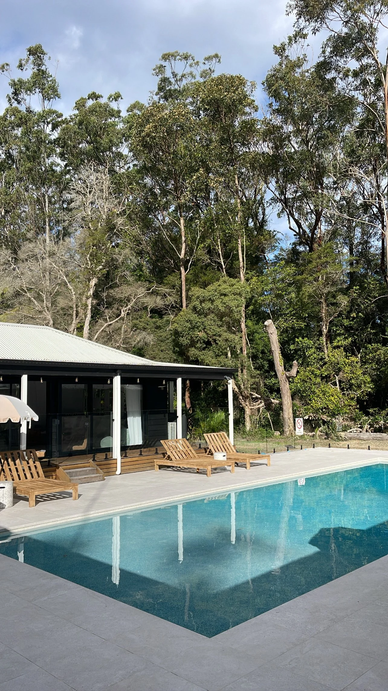 Swimming pool with lounge chairs and a building with a gray roof amid trees and a partly cloudy sky