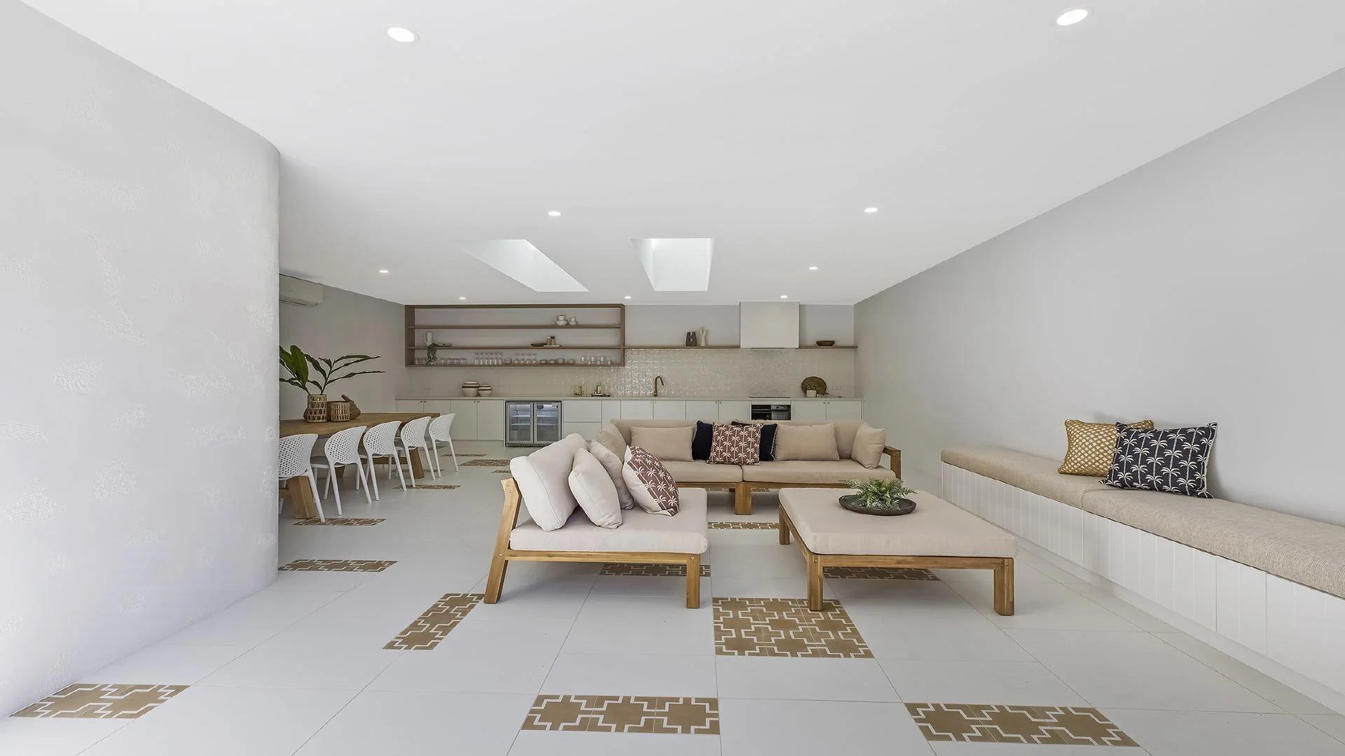 Bright and spacious modern living room with white walls, tiled floor with decorative brown accents, a large beige sectional sofa with throw pillows, a bench with pillows along the wall, a coffee table with a centerpiece, and an open kitchen and dining area in the background.