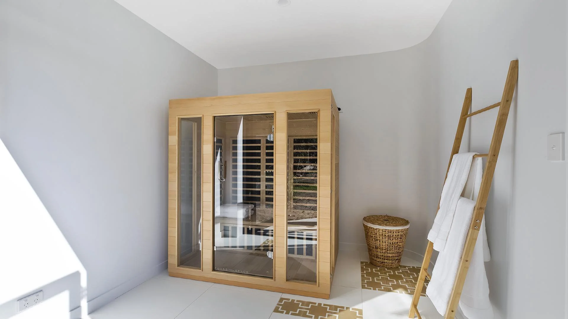 A home sauna with a glass door and wooden interior, next to a ladder with white towels and a wicker basket in a bright, white room.