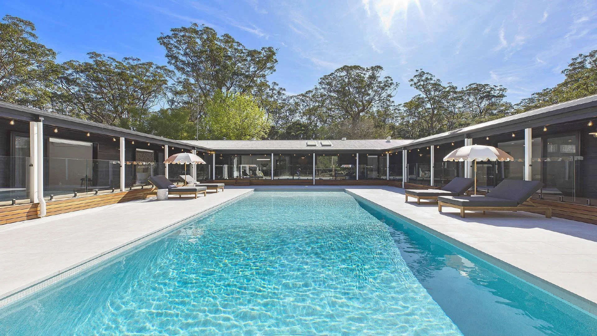A modern house with a swimming pool in the backyard, surrounded by lounge chairs, umbrellas, and trees.