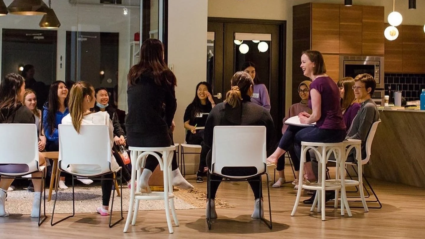 A group of women and girls participating in a discussion or workshop in a modern, cozy room, with some standing and others seated, smiling and engaging with each other.