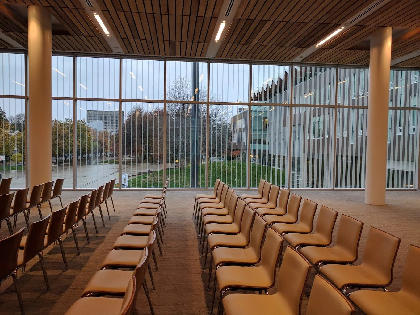 Conference room with beige chairs arranged in rows facing large floor-to-ceiling windows overlooking trees and a modern building.