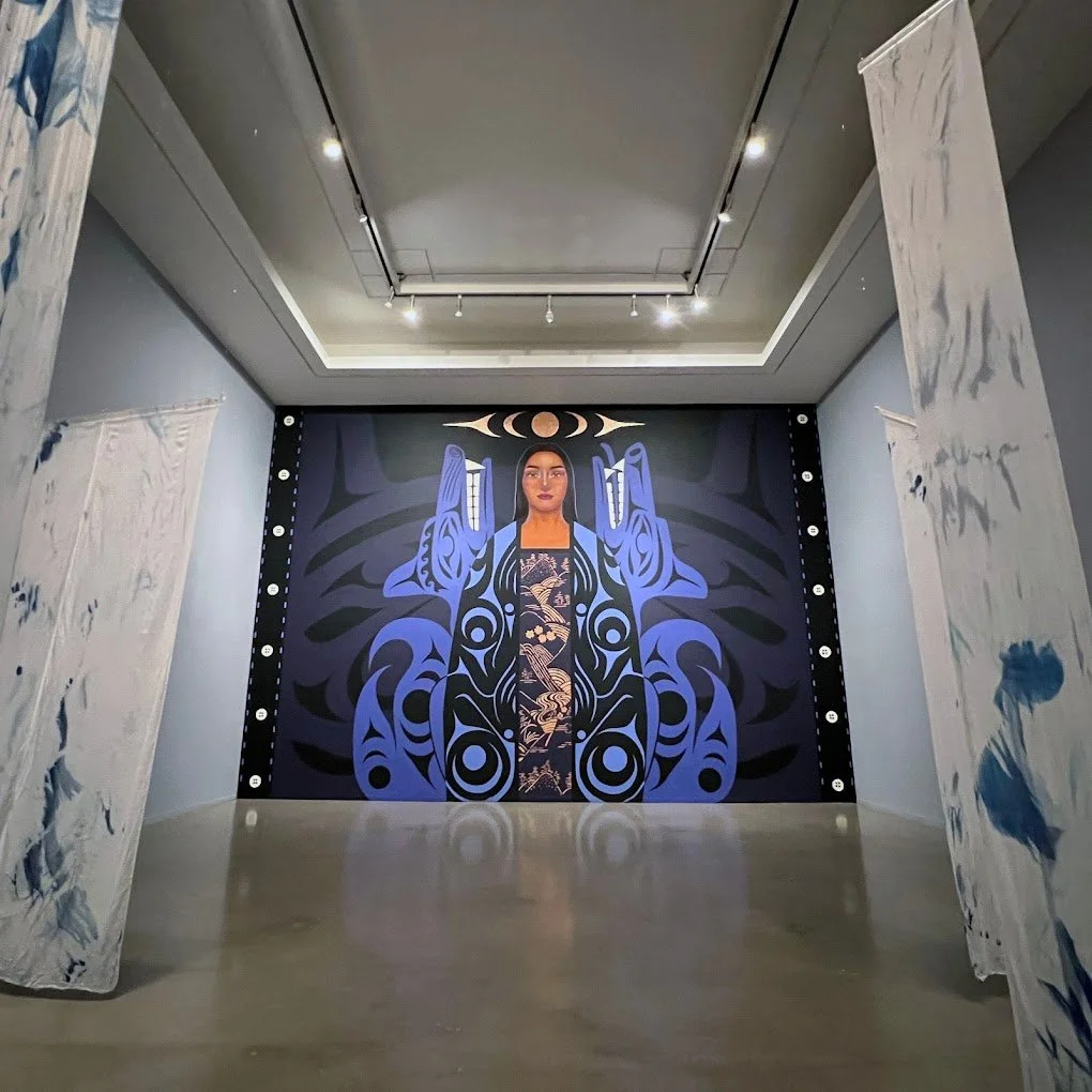 A large artwork featuring a woman with long dark hair, painted in blue tones with abstract patterns, framed by white fabric curtains with blue ink splatters on either side, in an art gallery setting.