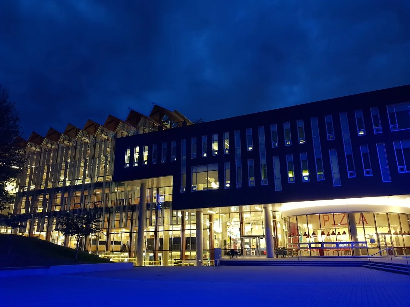 Modern multi-story building with glass exterior and illuminated interior, nighttime scene with a dark cloudy sky, a sign with the word 'PIZZA' inside.