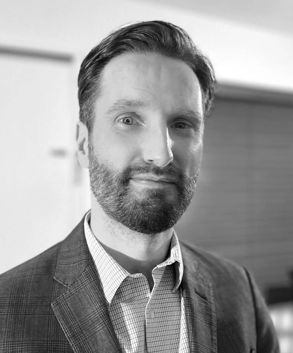 Black and white portrait of a man with a beard, wearing a plaid suit jacket and checkered shirt, in a professional setting.