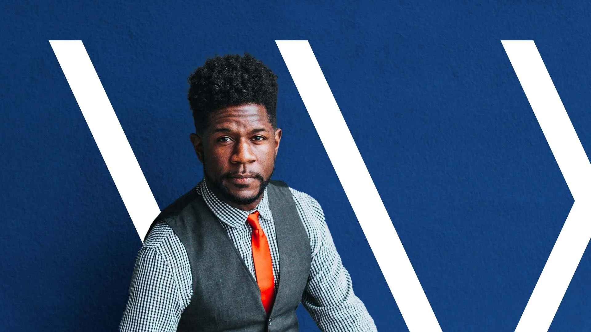 Man in a patterned shirt and vest with an orange tie against a blue background with white diagonal lines.