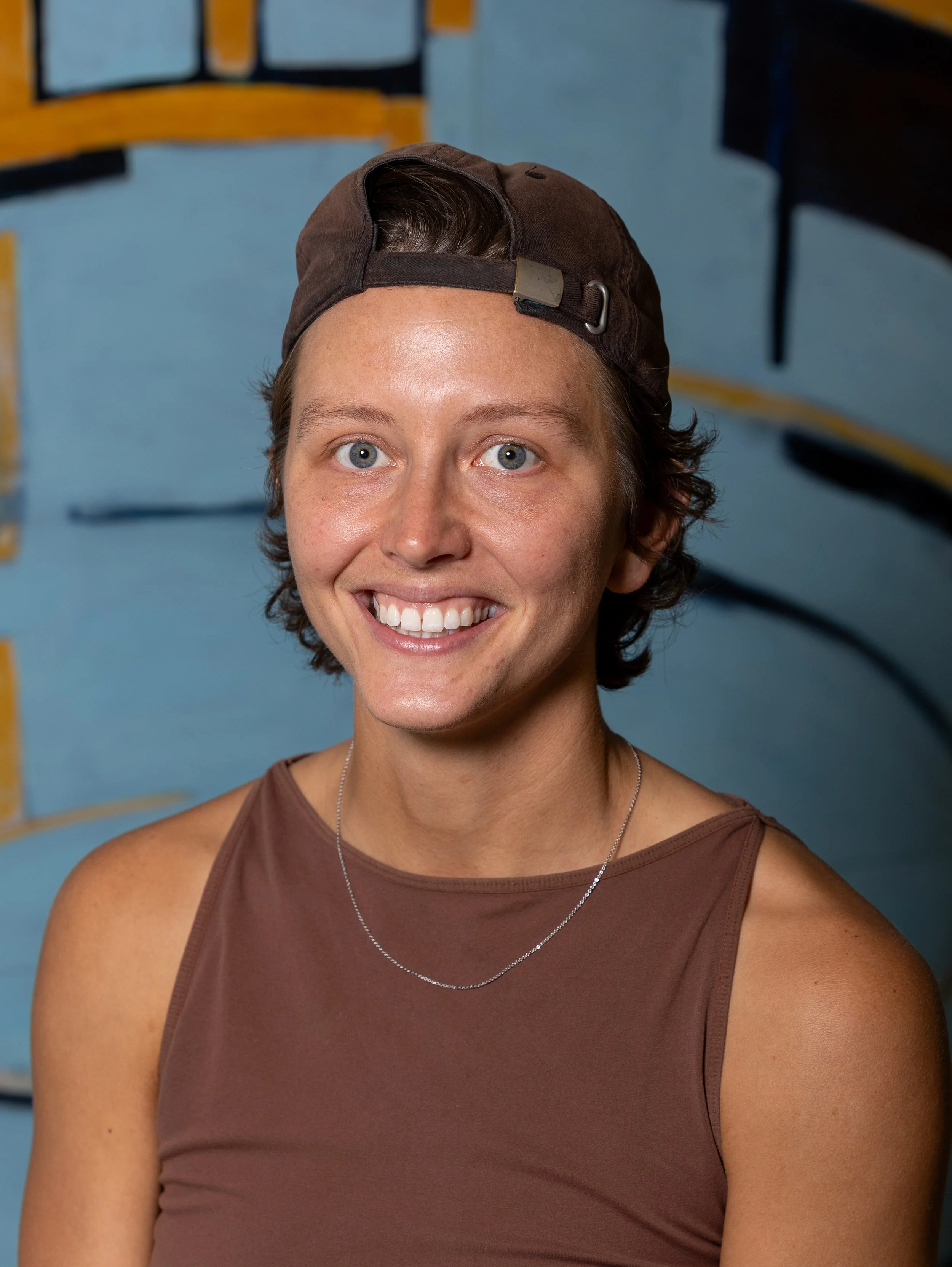 A person smiling warmly outdoors, wearing a black jacket, a striped neck gaiter, and a backwards dark baseball cap, with trees softly blurred in the background