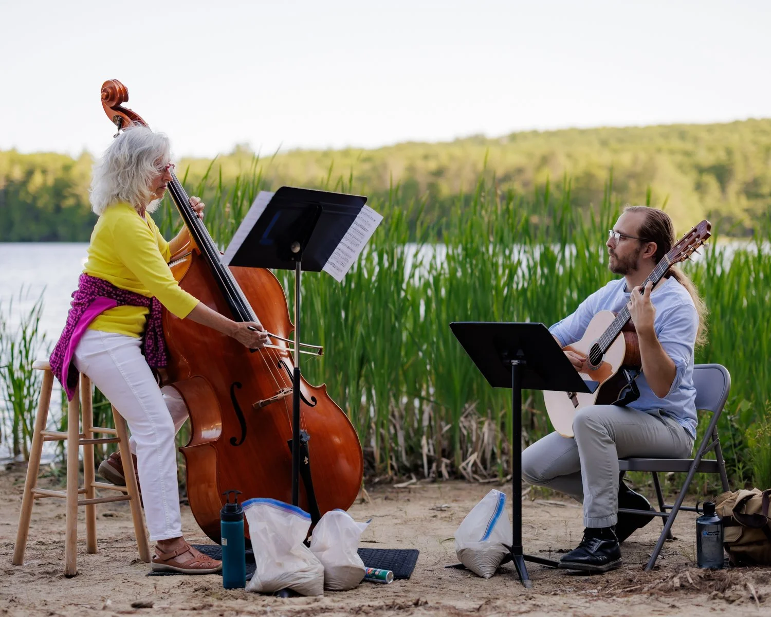An elderly woman and a young man play musical instruments outdoors near a lake with tall green reeds and trees in the background. The woman plays a double bass, and the man plays an acoustic guitar.