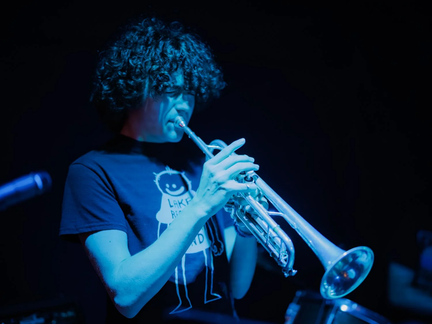 Person with curly hair playing trumpet on stage, wearing a black T-shirt with a smiley face and text, dark background.