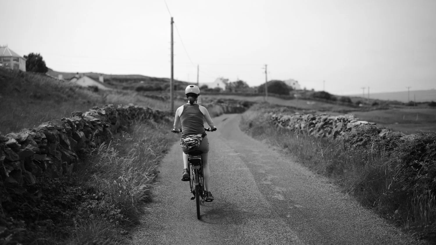 A woman biking on a rural gravel road with stone walls flanking both sides, hills and houses in the background, and utility poles lining the road.
