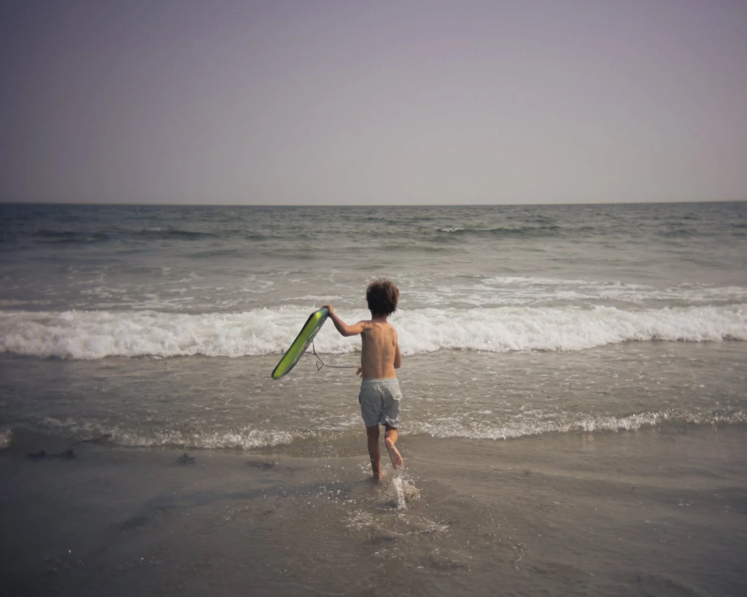 A young boy running into the ocean waves while holding a green and black boogie board.