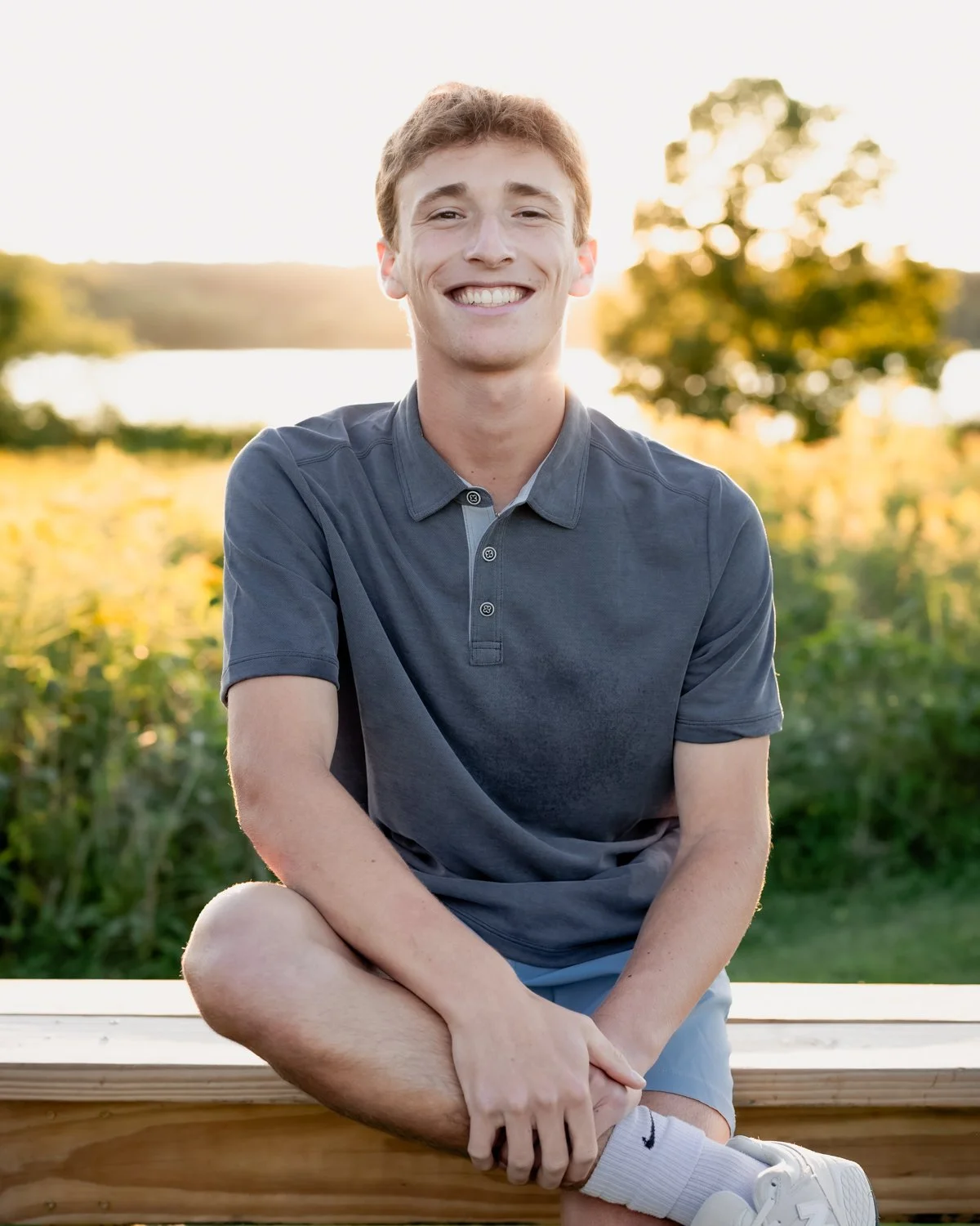 A young man with short red hair and a big smile sitting outdoors on a wooden bench with one leg crossed over the other. He's wearing a gray polo shirt, light blue shorts, white sneakers, and white socks. The background includes green trees and a body