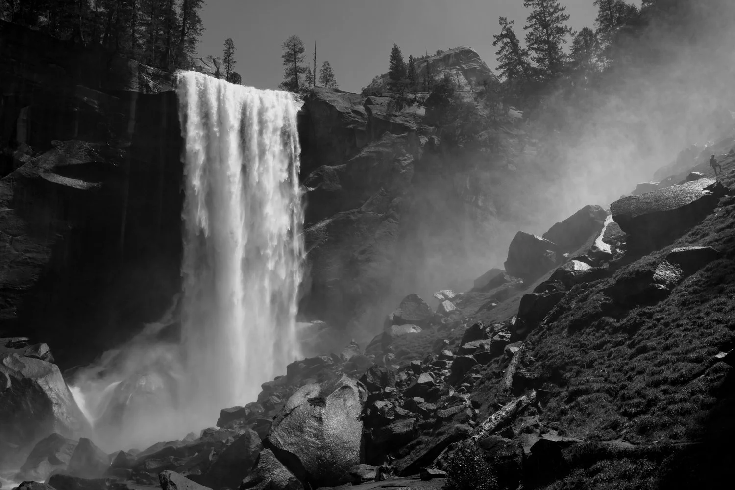 A black and white photograph of a large waterfall cascading down a rocky cliff into a misty pool below, surrounded by rocks and trees in a mountain landscape.
