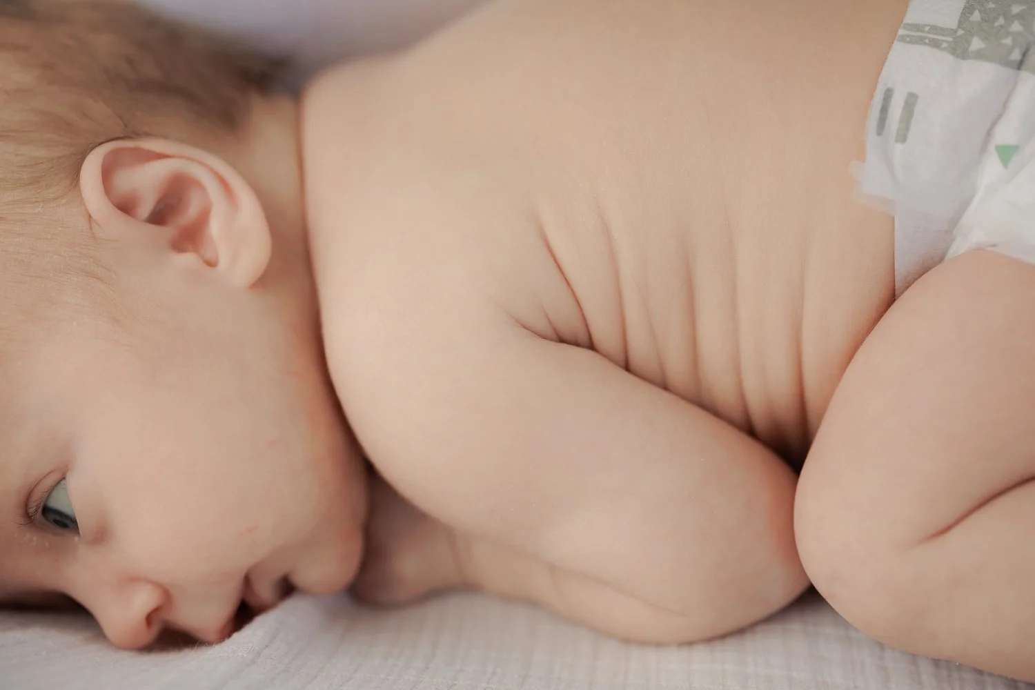 Close-up of a baby lying on its side, showing the baby's face, ear, and part of the body with skin folds, wearing a diaper.