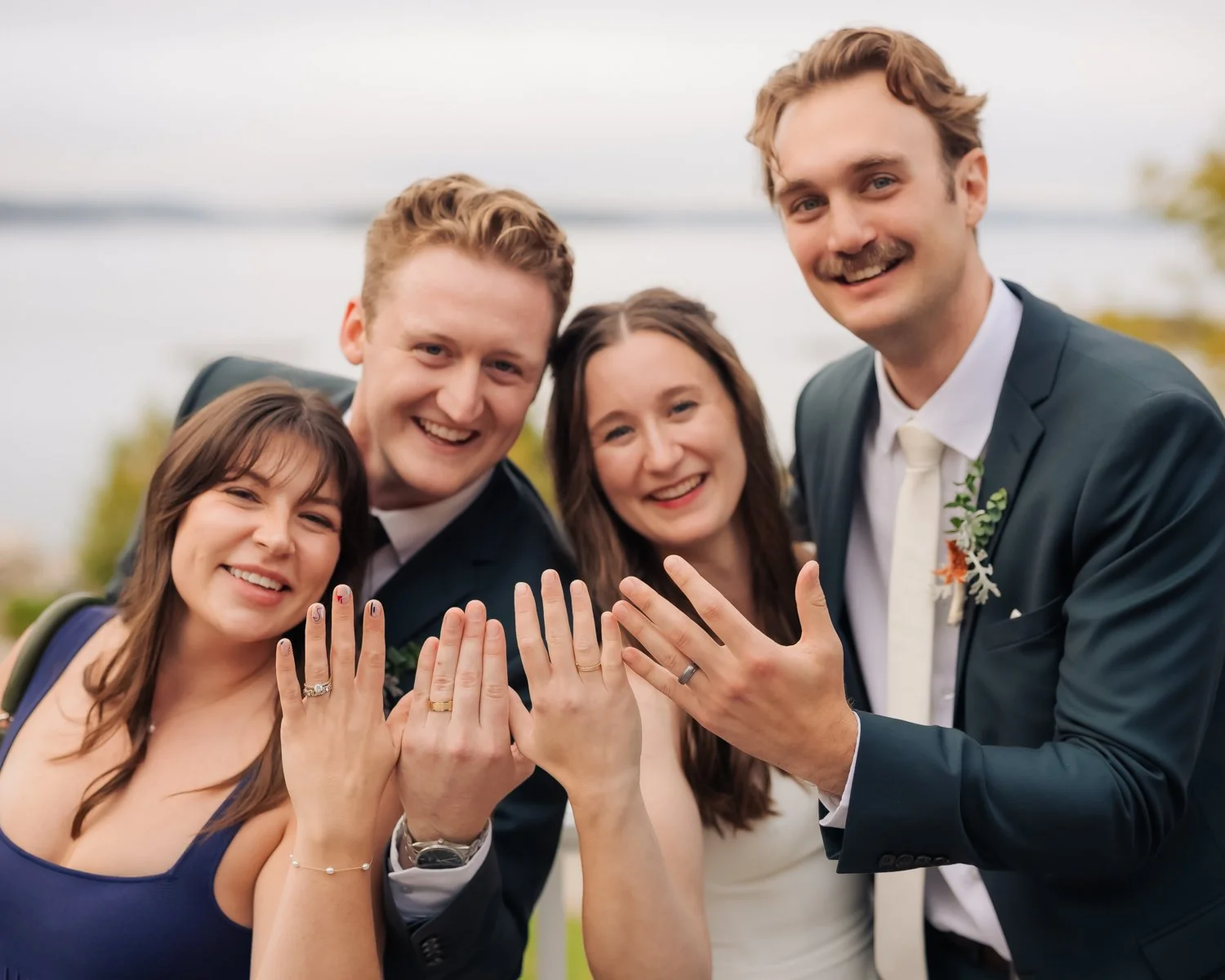 Group of four people showing off their wedding rings outdoors near a body of water, smiling at the camera.