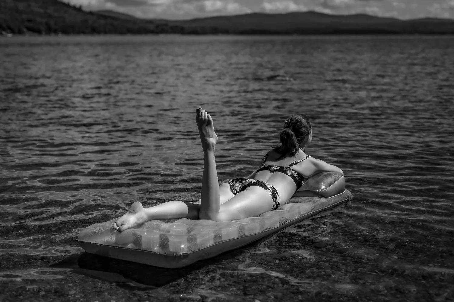 A woman lying on an inflatable raft in a body of water, wearing a swimsuit and holding her head with one arm, looking away from the camera.