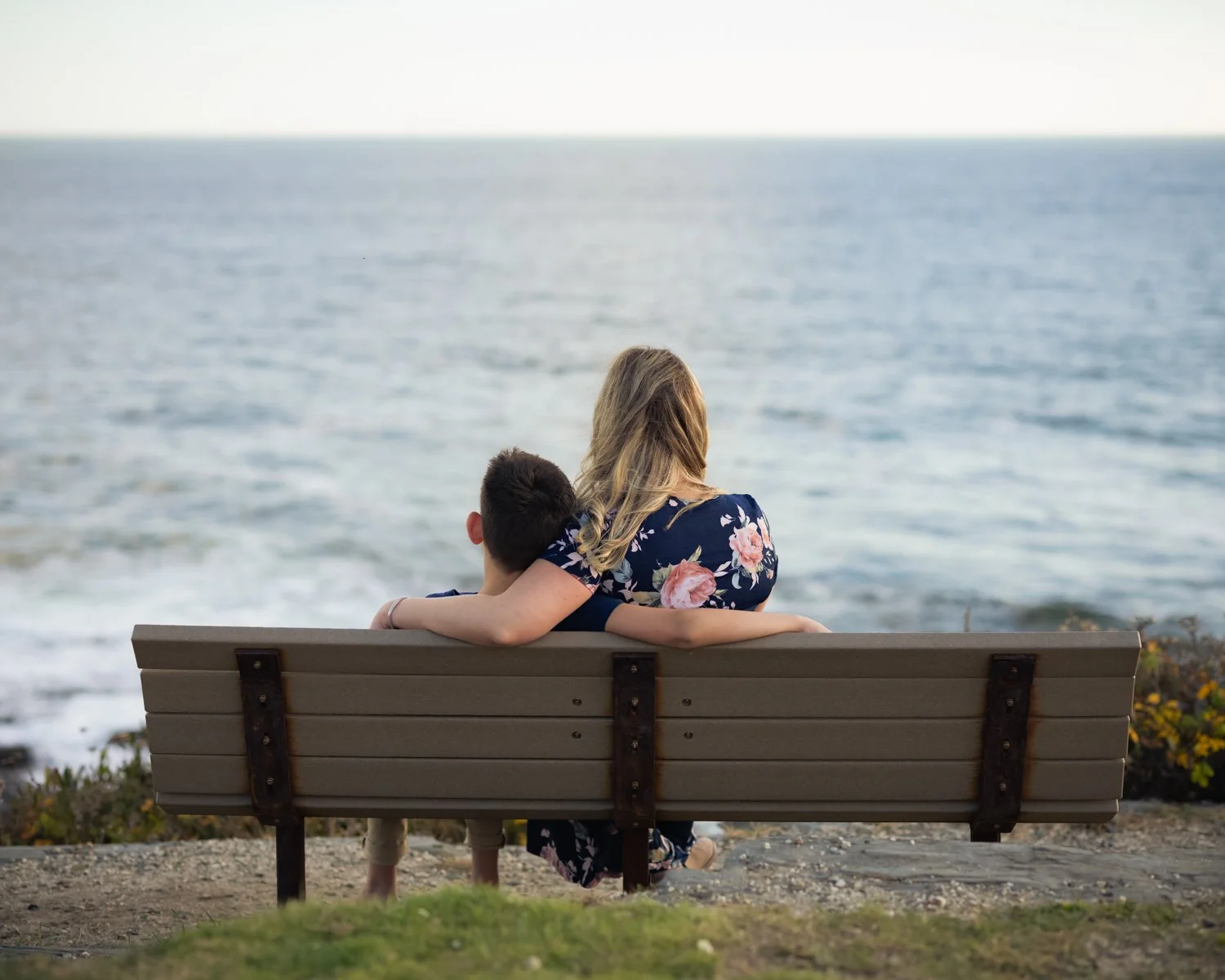 A woman and a boy sitting on a bench, looking at the ocean, with the woman resting her arm around the boy.