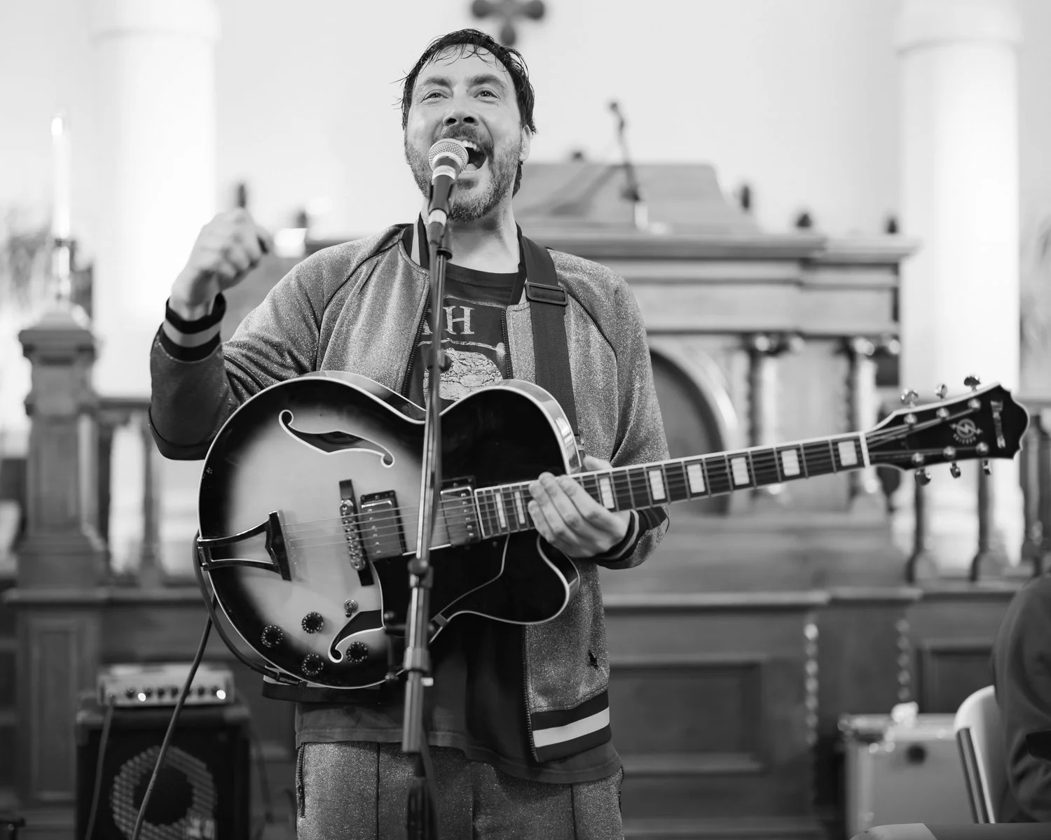 A man singing into a microphone while playing an electric guitar in a church or concert hall setting.