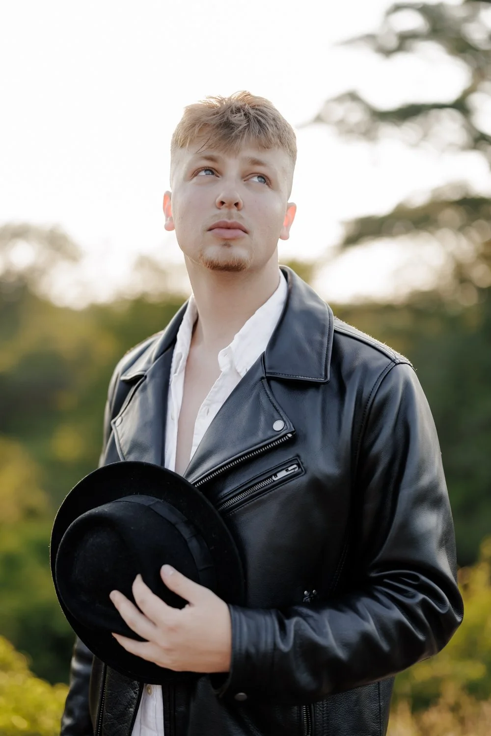 A young man with light brown hair, wearing a black leather jacket and white shirt, holding a black fedora hat, standing outdoors with trees blurred in the background during daylight.