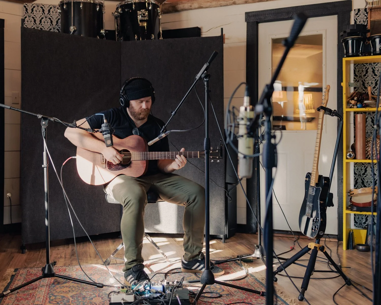A man wearing a black hat and black t-shirt is playing an acoustic guitar in a music studio. He is surrounded by microphones, microphones stands, and audio equipment. There is a black electric guitar on a stand and a yellow shelf with musical instrum