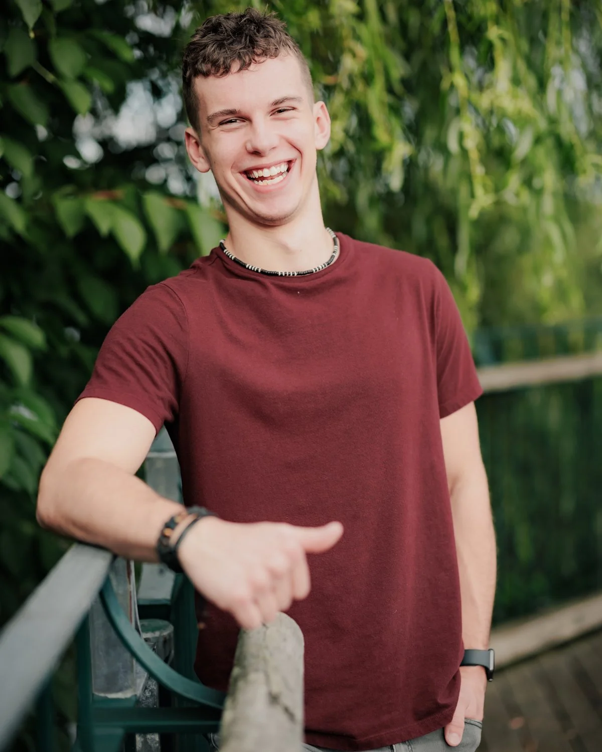 A young man with short, curly brown hair smiling and giving a thumbs-up while standing outdoors near a green fence and lush foliage.