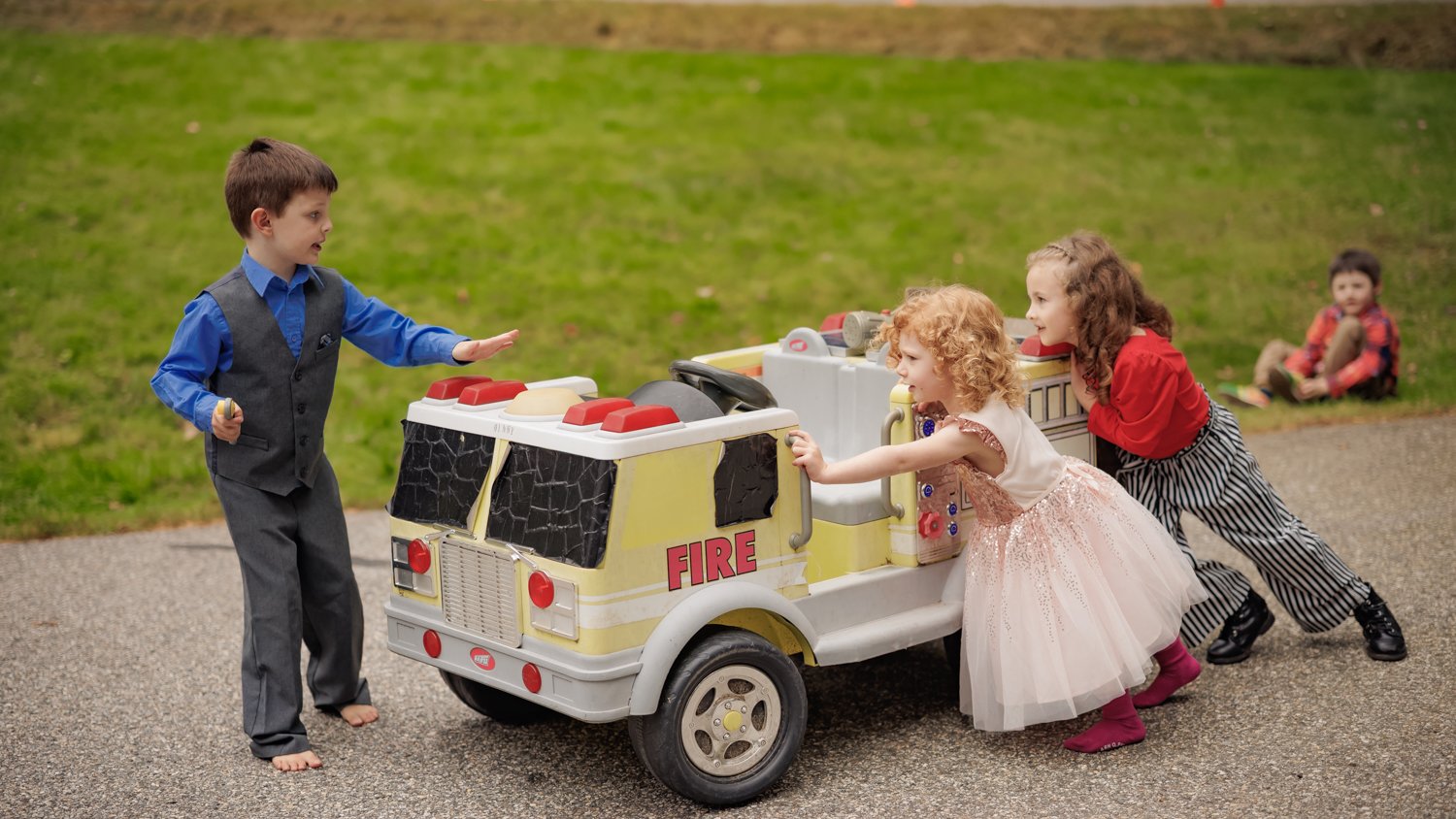 Children playing with a toy fire truck outdoors, with one child pretending to be a firefighter and others pushing the truck, two children sitting in the background.