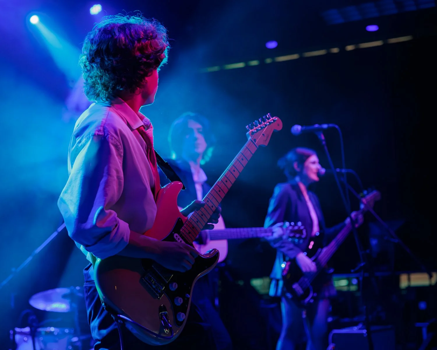 Three musicians playing guitars and singing on stage with colorful stage lighting.
