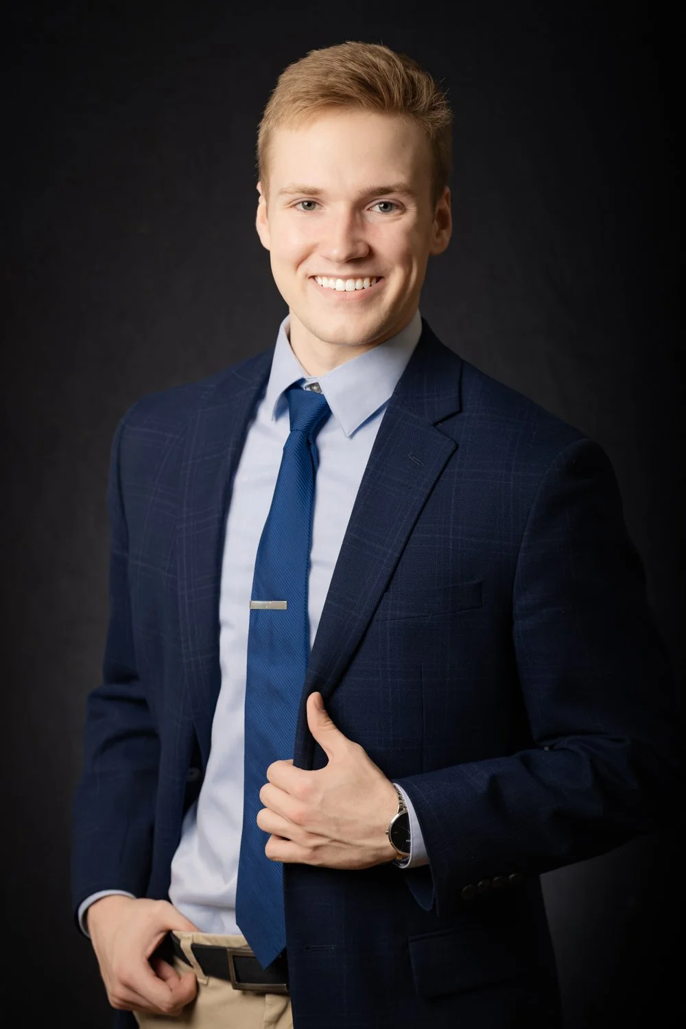 A young man with blonde hair smiling, dressed in a dark blue suit, light blue shirt, and matching blue tie, posed against a dark background.
