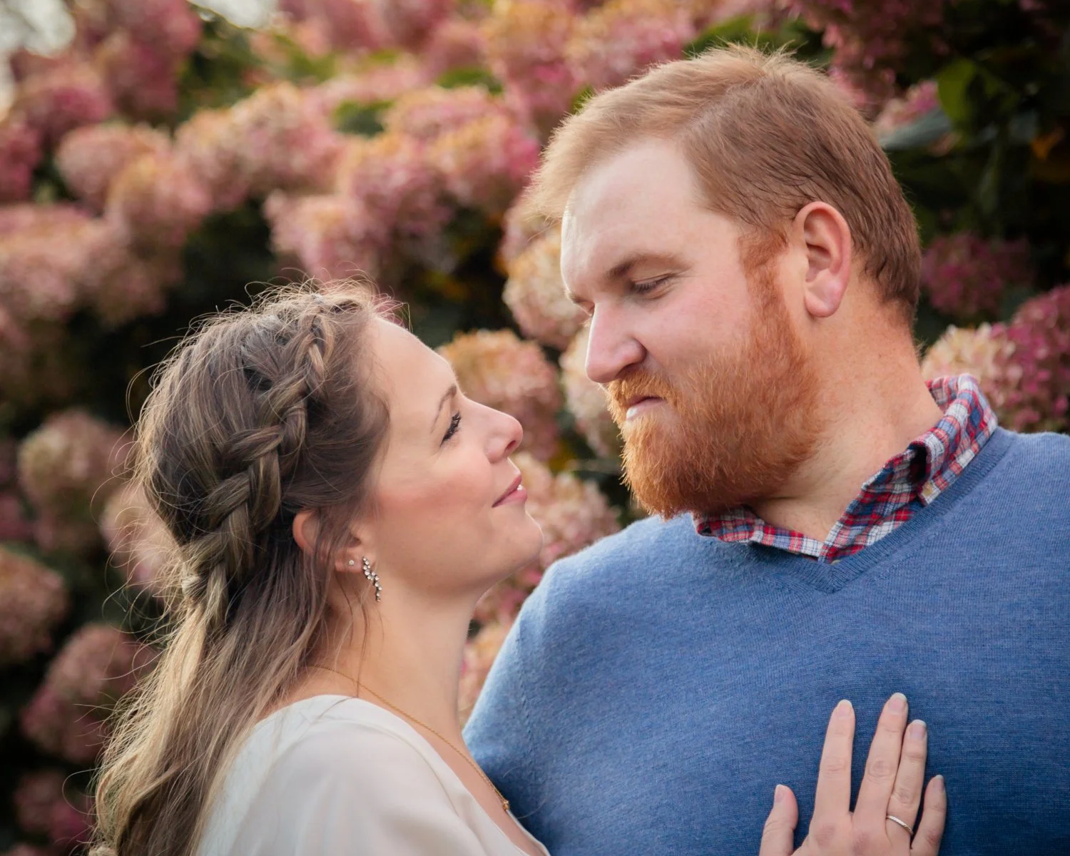 A couple standing close together outdoors in front of pink hydrangea flowers. The woman has long brown hair with a braid and is wearing a white top, and the man has red hair and a beard, wearing a blue sweater over a collared shirt. They are gazing i