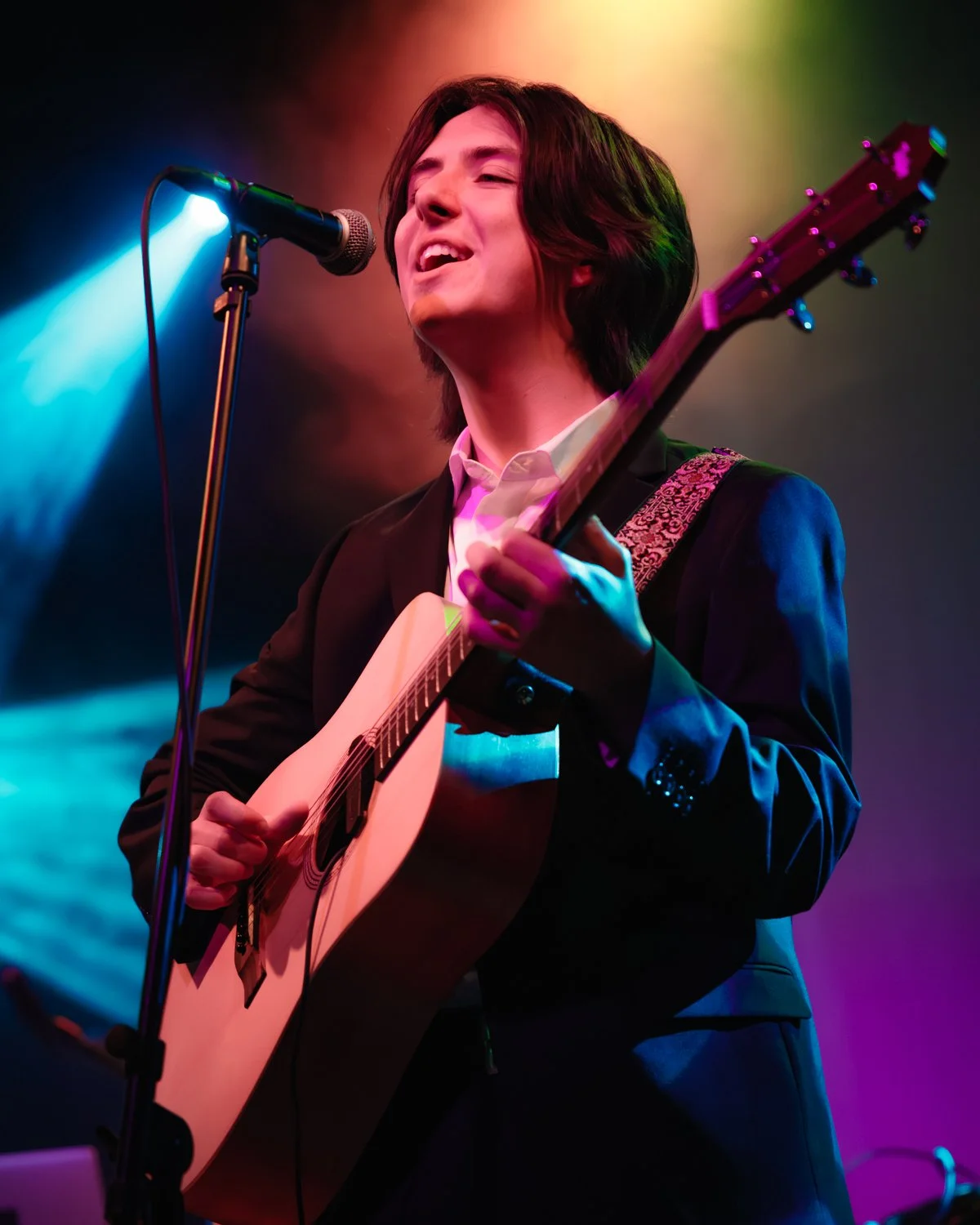 A person with shoulder-length hair singing into a microphone while playing an acoustic guitar on stage with colorful lighting.
