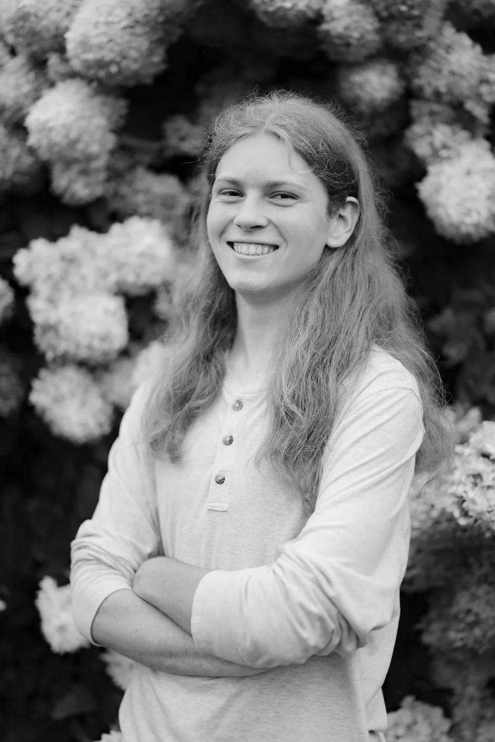 A young woman with long hair smiling, standing in front of a bush with large flowers.