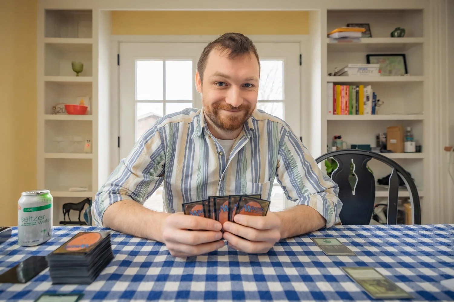 A man with a beard and striped shirt playing a card game at a table covered with a blue checkered tablecloth, with a soda can on the left and a few cards laid out on the table, in a well-lit room with shelves in the background.